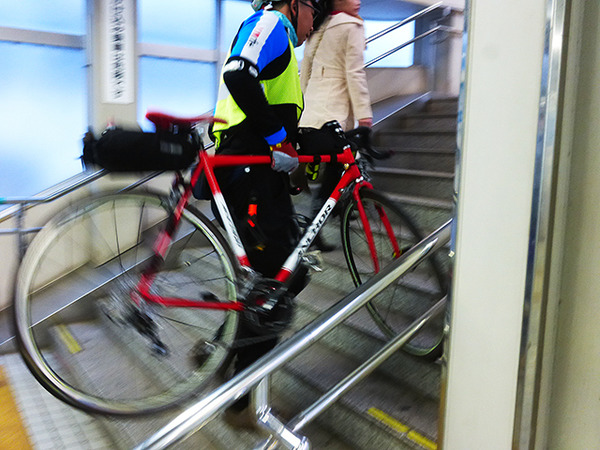 自転車を担いで階段を昇り、モノレール乗り場へと向かう参加者たち（11月30日、千葉モノレール実証実験「サイクル＆モノレール」）
